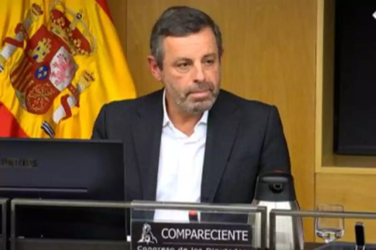 Hombre de mediana edad con barba sentado en una sala oficial con una bandera de España al fondo y un cartel que dice compareciente
