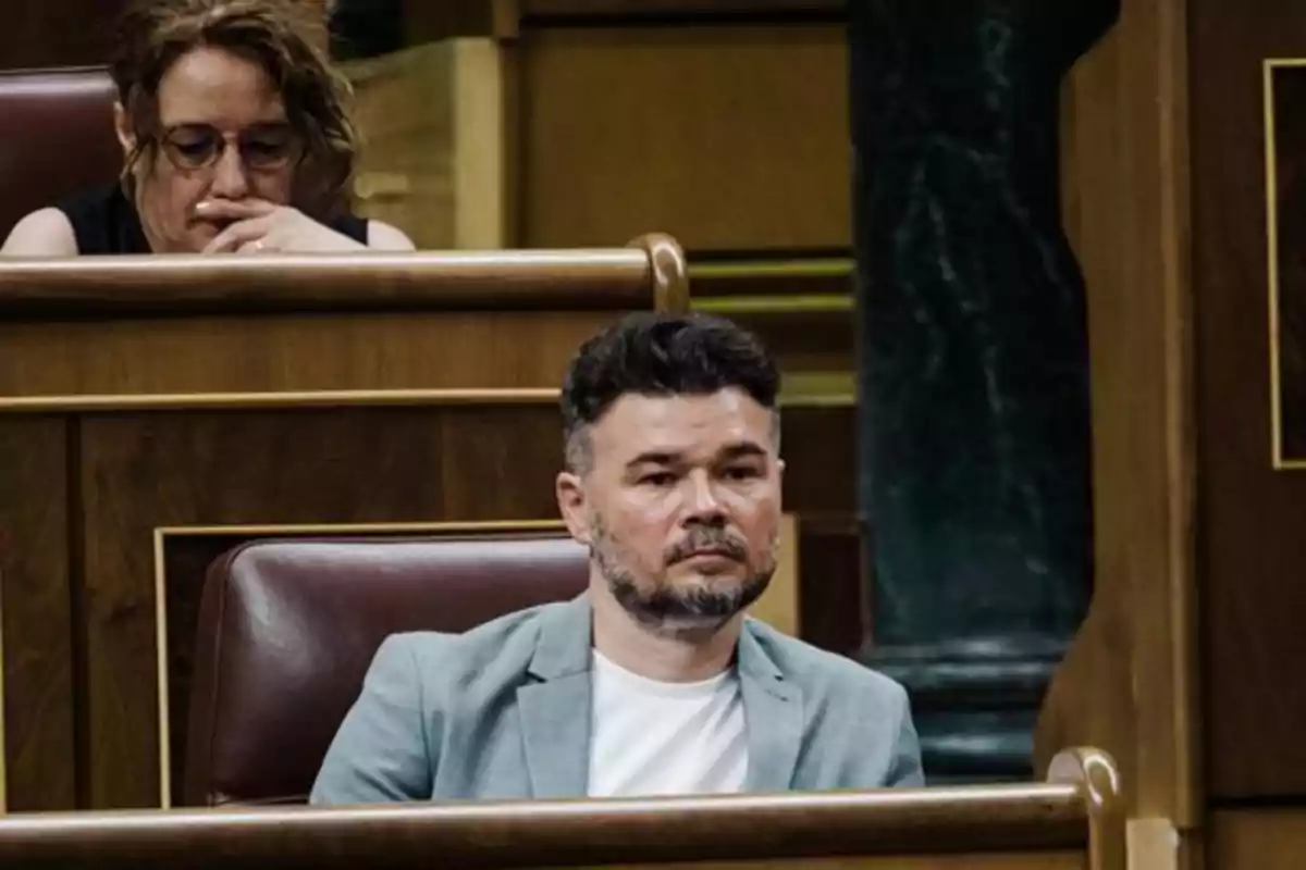 Hombre sentado en un escaño de madera con expresión seria y una mujer al fondo pensativa Hombre sentado en un escaño de madera con expresión seria y una mujer al fondo pensativa