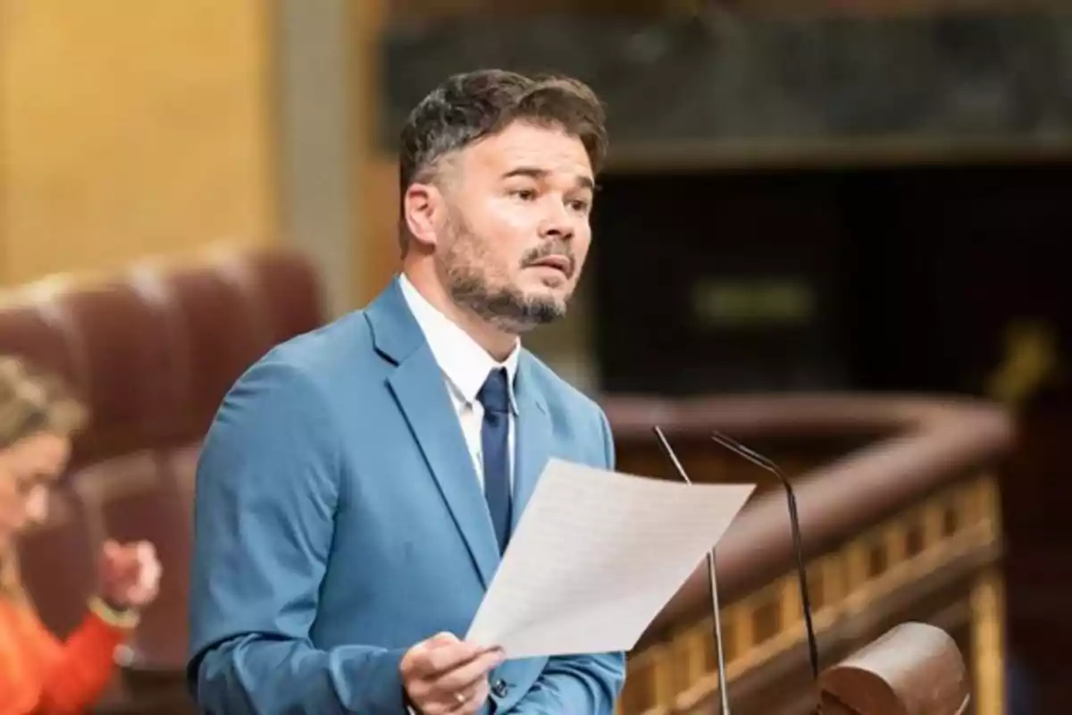 Hombre con barba y traje azul hablando en un podio mientras sostiene una hoja de papel en un entorno parlamentario