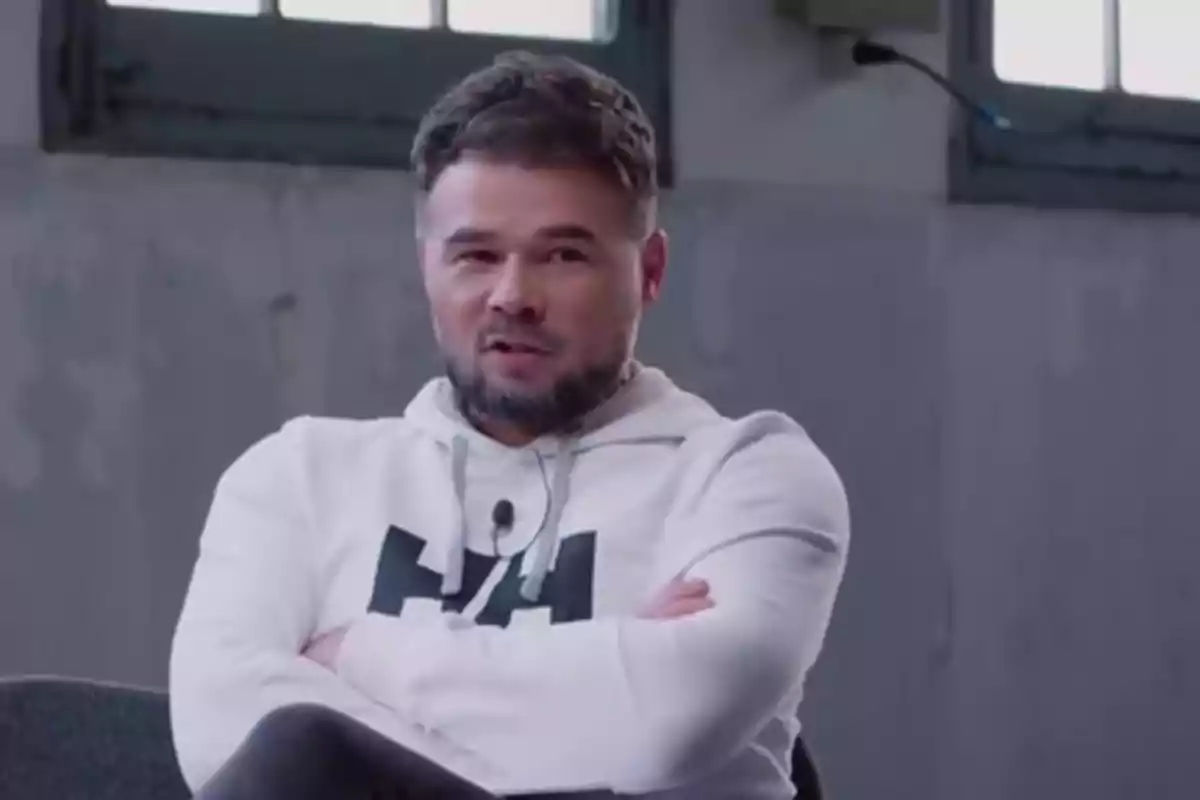Hombre con barba y cabello corto usando sudadera blanca sentado con los brazos cruzados en un ambiente interior Hombre con barba y cabello corto usando sudadera blanca sentado con los brazos cruzados en un ambiente interior