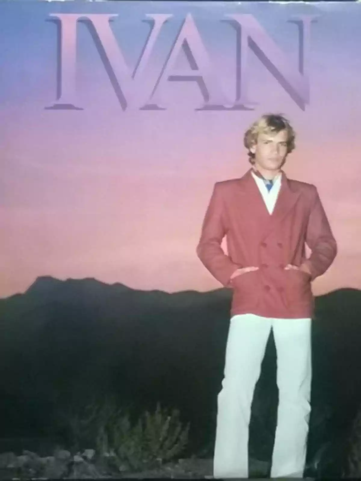 Hombre rubio con saco rojo y pantalón blanco posando frente a un paisaje montañoso al atardecer con el texto IVAN en la parte superior Hombre rubio con saco rojo y pantalón blanco posando frente a un paisaje montañoso al atardecer con el texto IVAN en la parte superior
