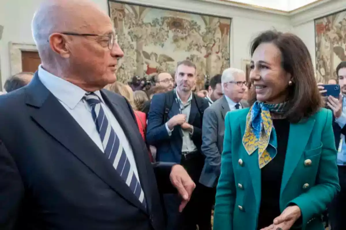 Dos personas elegantemente vestidas conversan y sonríen en un evento social con varias personas al fondo en una sala decorada con tapices.