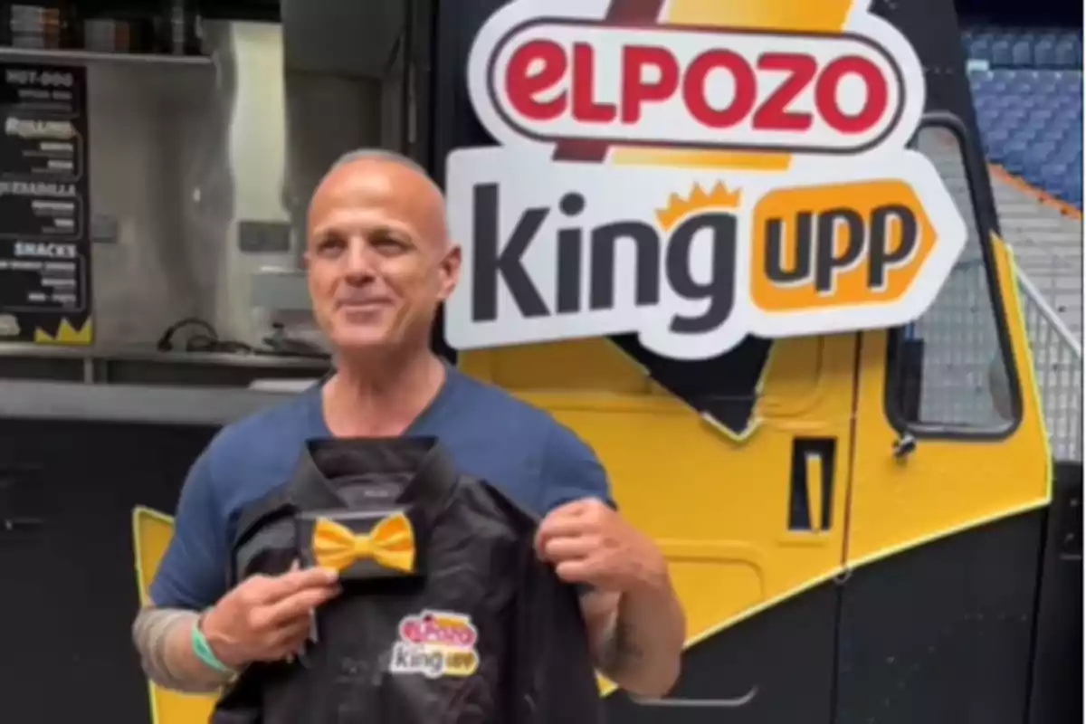 Hombre sonriente sostiene una camisa negra con un moño amarillo frente a un camión de comida con el logo de El Pozo King Upp Hombre sonriente sostiene una camisa negra con un moño amarillo frente a un camión de comida con el logo de El Pozo King Upp