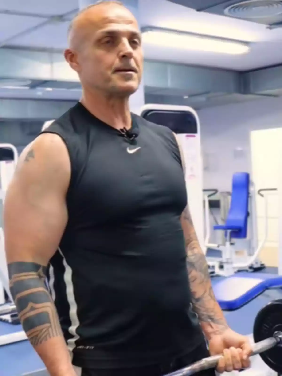 Hombre musculoso con tatuajes en los brazos usando camiseta negra sin mangas mientras sostiene una barra en un gimnasio moderno Hombre musculoso con tatuajes en los brazos usando camiseta negra sin mangas mientras sostiene una barra en un gimnasio moderno
