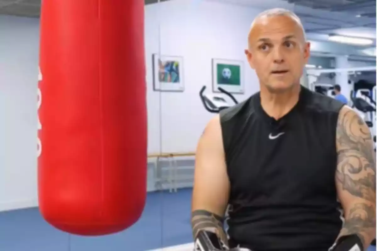 Hombre con camiseta sin mangas y tatuajes en el brazo sentado en un gimnasio junto a un saco de boxeo rojo Hombre con camiseta sin mangas y tatuajes en el brazo sentado en un gimnasio junto a un saco de boxeo rojo