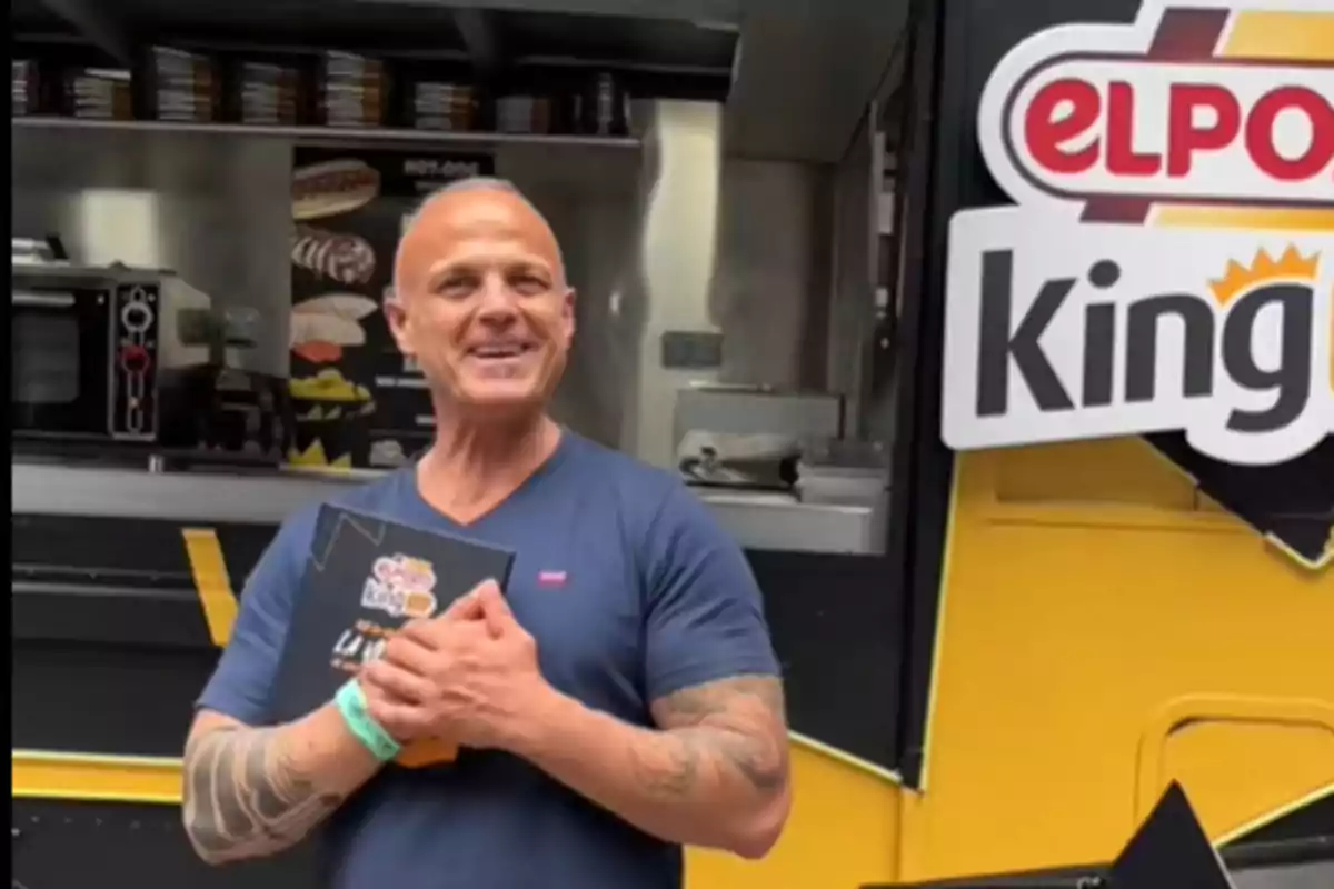 Hombre sonriente con tatuajes en los brazos sostiene un menú frente a un food truck amarillo con el logo de ElPozo King Hombre sonriente con tatuajes en los brazos sostiene un menú frente a un food truck amarillo con el logo de ElPozo King