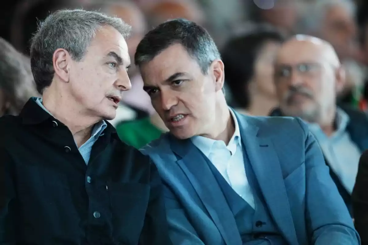 Dos hombres de mediana edad conversan de cerca en un evento formal rodeados de otras personas desenfocadas al fondo Dos hombres de mediana edad conversan de cerca en un evento formal rodeados de otras personas desenfocadas al fondo