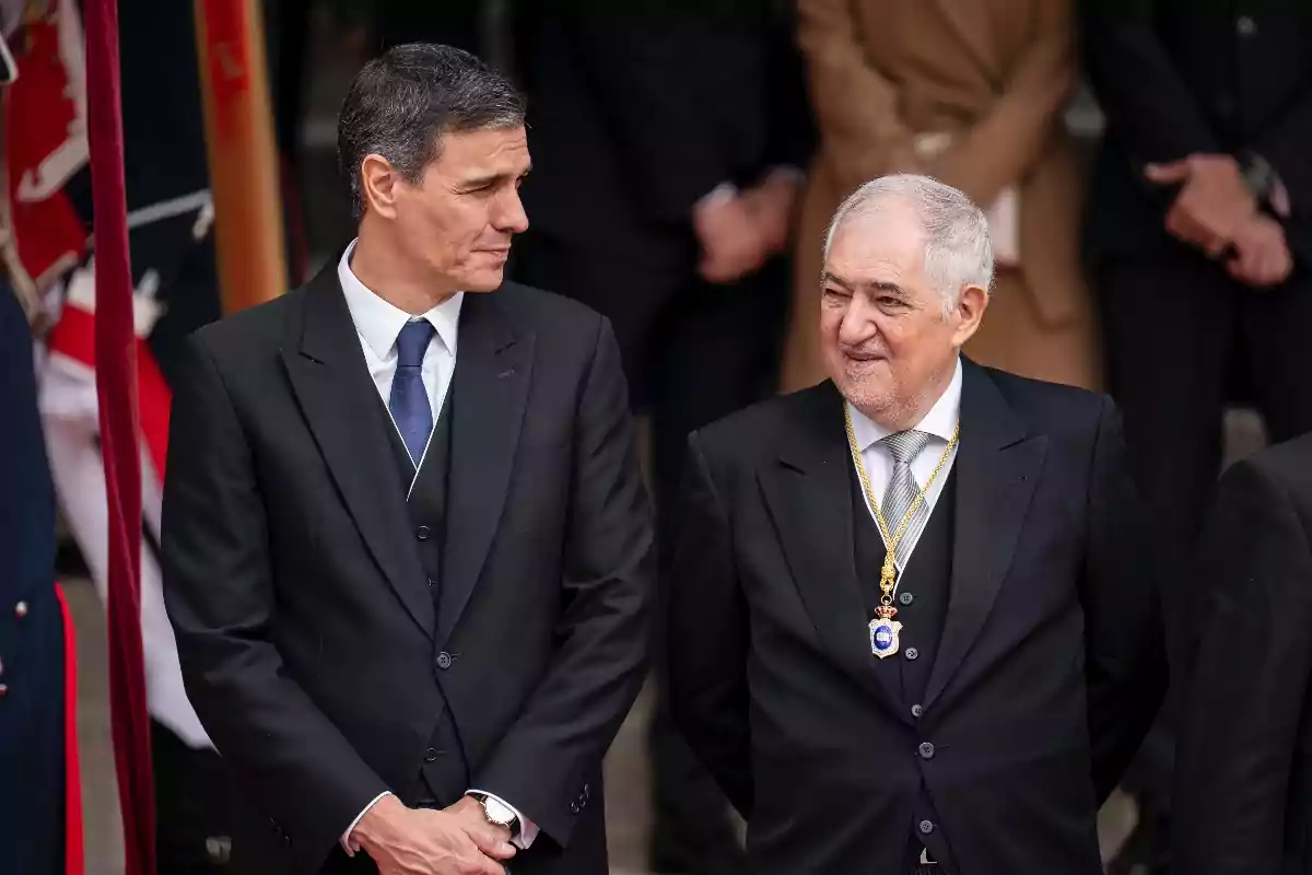 Dos hombres vestidos de traje oscuro conversan en un evento formal, uno de ellos lleva una medalla dorada colgada al cuello.