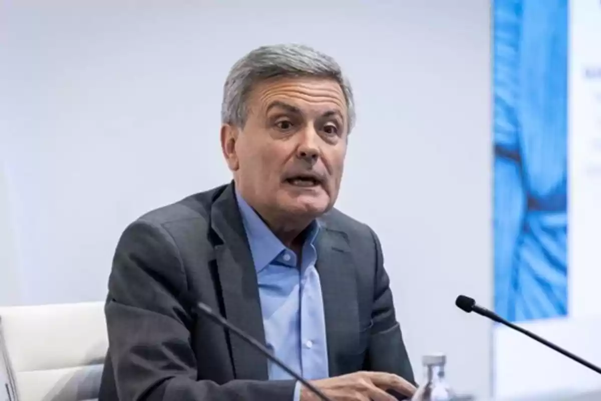 Hombre de cabello canoso con traje gris y camisa azul hablando frente a un micrófono en una sala de conferencias Hombre de cabello canoso con traje gris y camisa azul hablando frente a un micrófono en una sala de conferencias