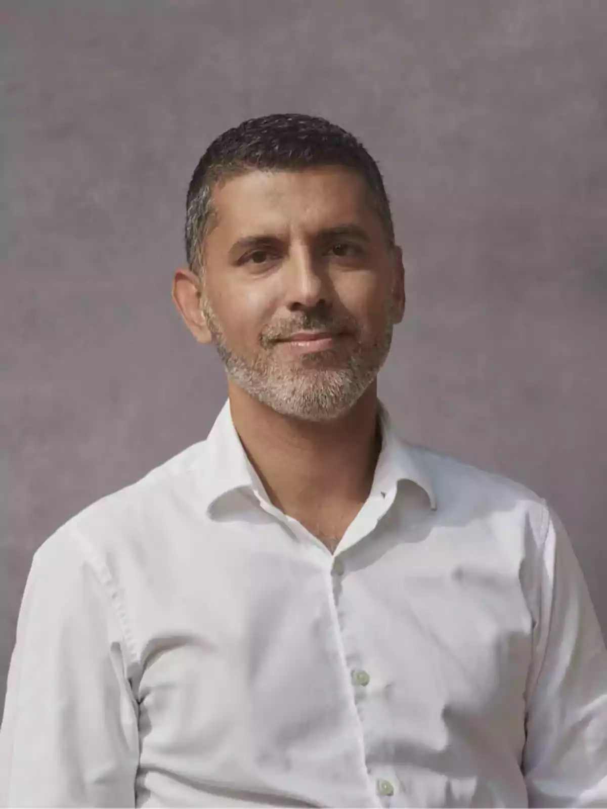 Hombre de cabello corto y barba canosa vistiendo camisa blanca frente a un fondo gris Hombre de cabello corto y barba canosa vistiendo camisa blanca frente a un fondo gris