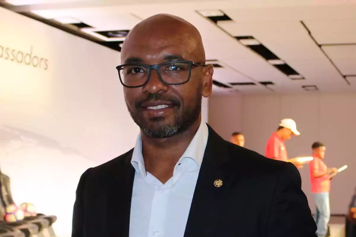 Hombre con gafas y barba vestido con traje oscuro y camisa blanca en un evento en interiores con personas al fondo
