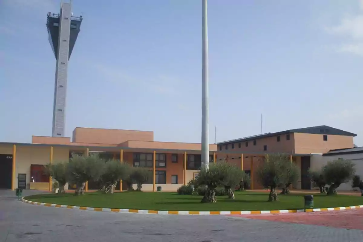 Edificio moderno de ladrillo con una torre alta al fondo, rodeado de césped y varios olivos en el exterior Edificio moderno de ladrillo con una torre alta al fondo, rodeado de césped y varios olivos en el exterior