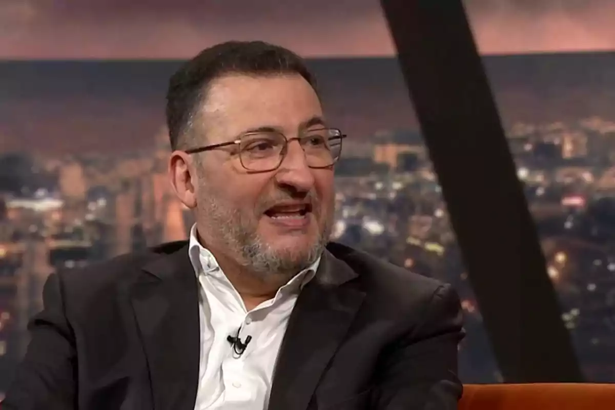 Hombre con gafas y barba hablando en un programa de televisión con fondo de ciudad desenfocado Hombre con gafas y barba hablando en un programa de televisión con fondo de ciudad desenfocado