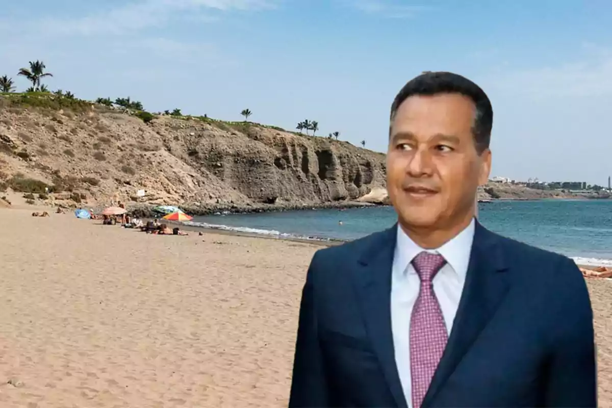 Hombre con traje y corbata de pie en una playa con acantilados y sombrillas al fondo