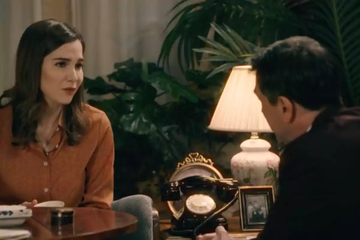 Begoña y Gabriel conversan sentados en una sala decorada con plantas, una lámpara de mesa, un teléfono antiguo y una fotografía enmarcada. Captura de 'Sueños de libertad' Begoña y Gabriel conversan sentados en una sala decorada con plantas, una lámpara de mesa, un teléfono antiguo y una fotografía enmarcada. Captura de 'Sueños de libertad'