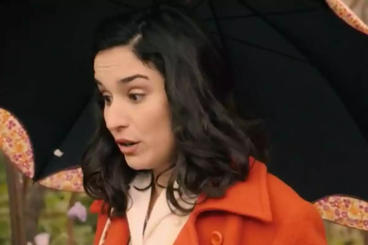 Cristina de cabello oscuro con expresión de sorpresa usando un abrigo naranja y sosteniendo un paraguas negro con borde decorado. Captura de 'Sueños de libertad' Cristina de cabello oscuro con expresión de sorpresa usando un abrigo naranja y sosteniendo un paraguas negro con borde decorado. Captura de 'Sueños de libertad'