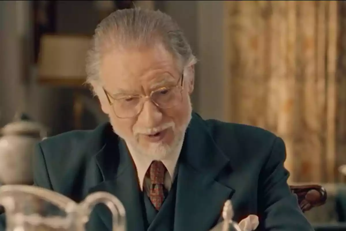 Damián de cabello canoso y barba blanca usando gafas y traje oscuro sentado en una mesa en un ambiente elegante. Captura de 'Sueños de libertad' Damián de cabello canoso y barba blanca usando gafas y traje oscuro sentado en una mesa en un ambiente elegante. Captura de 'Sueños de libertad'