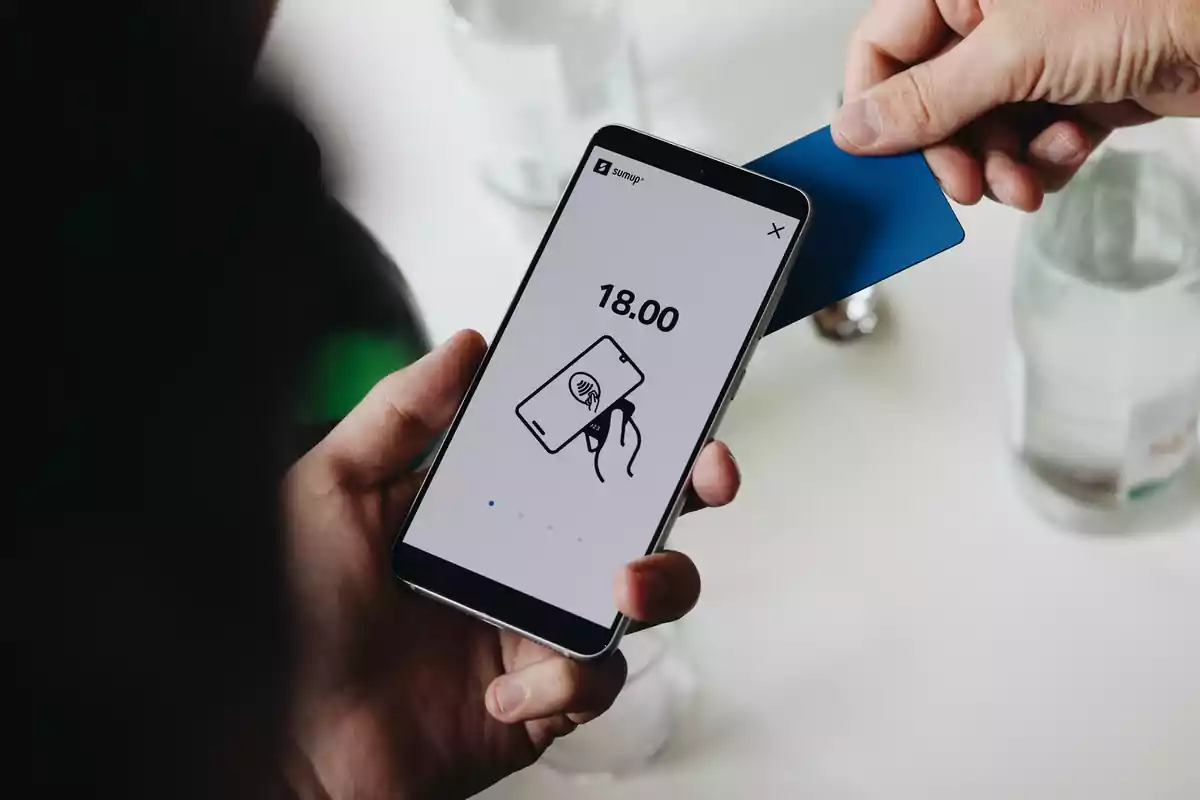 Persona sosteniendo un teléfono móvil que muestra una pantalla de pago de 18.00 mientras otra persona acerca una tarjeta azul para realizar el pago electrónico Persona sosteniendo un teléfono móvil que muestra una pantalla de pago de 18.00 mientras otra persona acerca una tarjeta azul para realizar el pago electrónico
