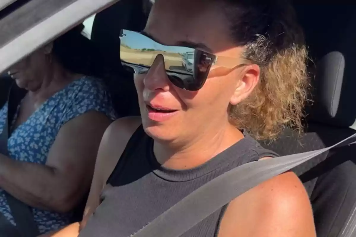 Tamara con gafas de sol grandes y camiseta sin mangas negras sentada en el asiento del copiloto de un coche con el cinturón de seguridad puesto y otra persona al volante Tamara con gafas de sol grandes y camiseta sin mangas negras sentada en el asiento del copiloto de un coche con el cinturón de seguridad puesto y otra persona al volante