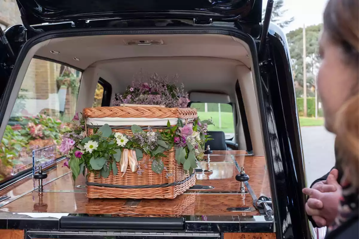 Ataúd de mimbre decorado con flores en la parte trasera de un coche fúnebre mientras una persona observa Ataúd de mimbre decorado con flores en la parte trasera de un coche fúnebre mientras una persona observa