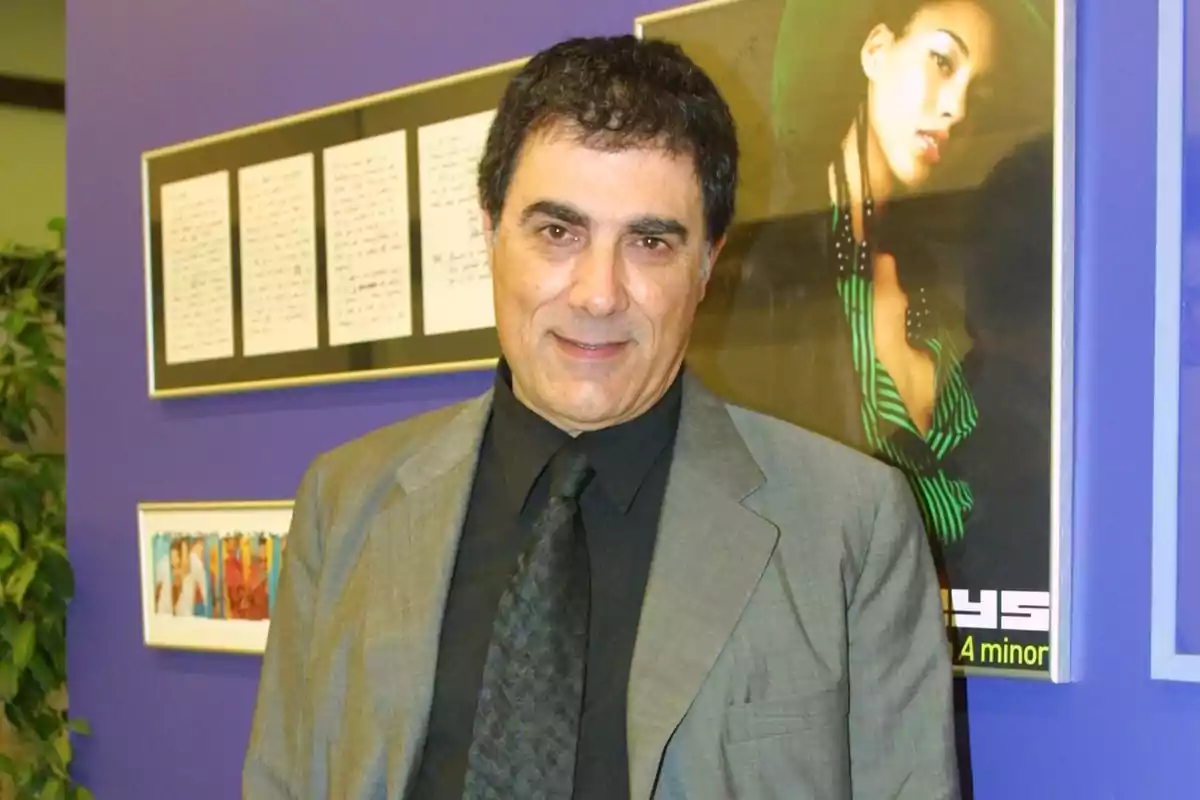 Hombre de cabello oscuro con traje gris y camisa negra posando frente a una pared morada decorada con cuadros y una fotografía de una mujer