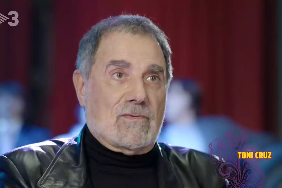 Hombre de cabello canoso y barba corta con chaqueta de cuero negra y suéter oscuro, fondo desenfocado con cortinas rojas y el nombre Toni Cruz en la esquina inferior derecha Hombre de cabello canoso y barba corta con chaqueta de cuero negra y suéter oscuro, fondo desenfocado con cortinas rojas y el nombre Toni Cruz en la esquina inferior derecha