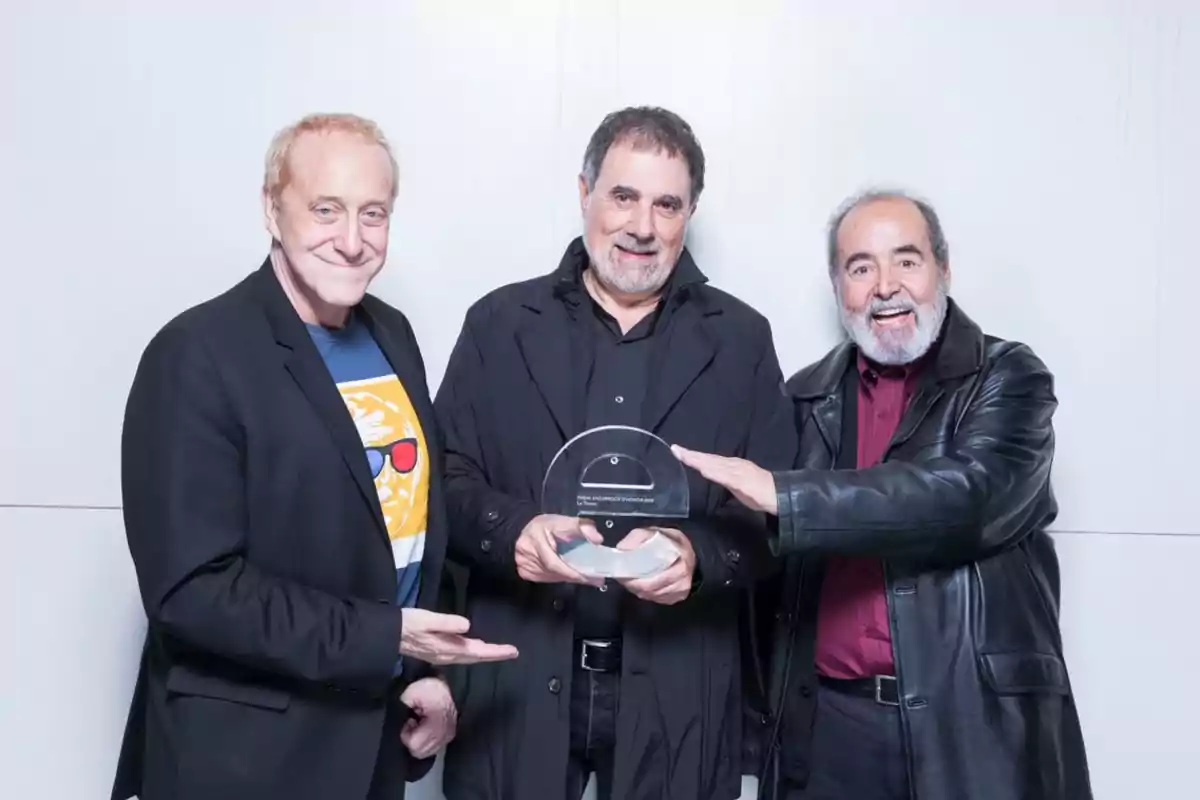 Tres hombres sonríen mientras uno de ellos sostiene un trofeo transparente y los otros dos lo señalan, todos vestidos con ropa oscura frente a una pared blanca Tres hombres sonríen mientras uno de ellos sostiene un trofeo transparente y los otros dos lo señalan, todos vestidos con ropa oscura frente a una pared blanca