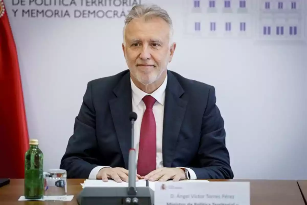 Hombre de cabello canoso y barba sentado en una mesa de conferencia con micrófono, usando traje oscuro y corbata roja, con una botella de agua y un vaso frente a él