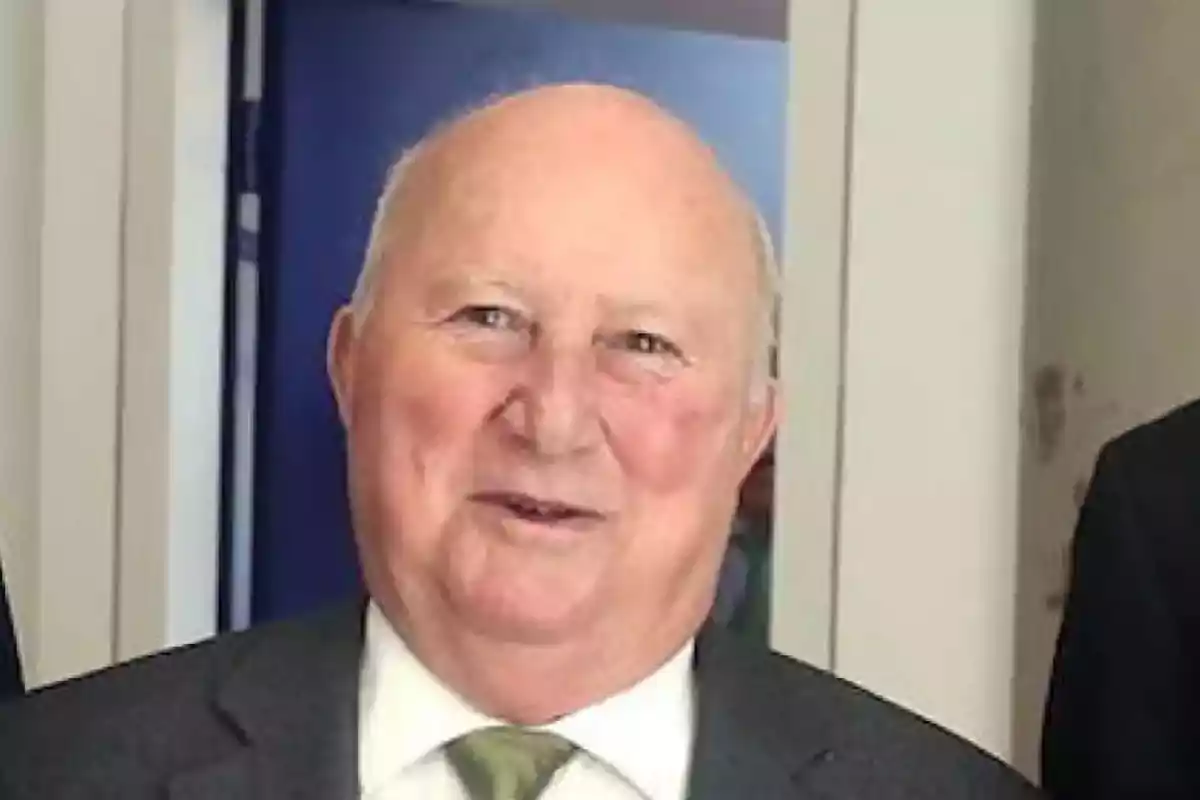Hombre mayor calvo con traje y corbata verde sonriendo frente a una pared blanca y fondo azul