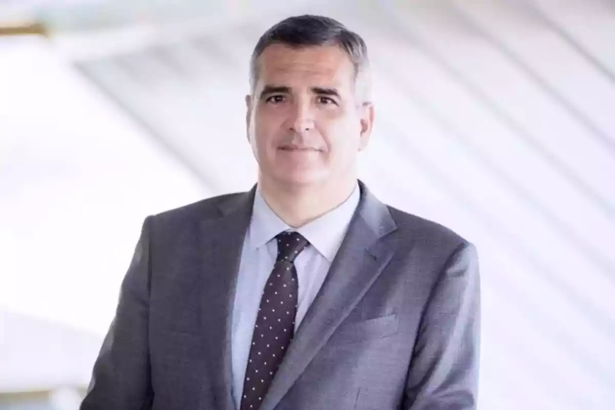 Hombre de mediana edad con cabello corto y canoso vistiendo un traje gris y corbata oscura con puntos blancos en un entorno moderno y luminoso Hombre de mediana edad con cabello corto y canoso vistiendo un traje gris y corbata oscura con puntos blancos en un entorno moderno y luminoso