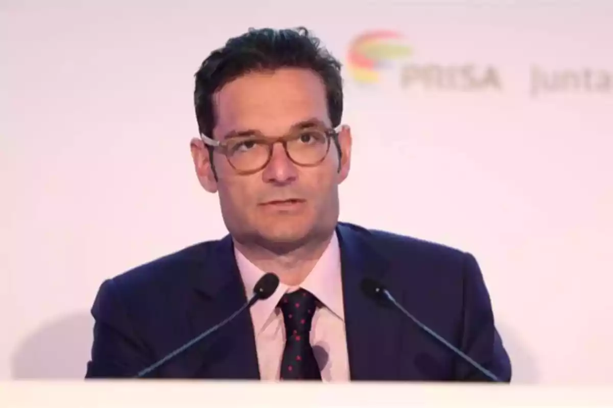 Hombre con gafas y traje hablando frente a dos micrófonos en un evento profesional Hombre con gafas y traje hablando frente a dos micrófonos en un evento profesional