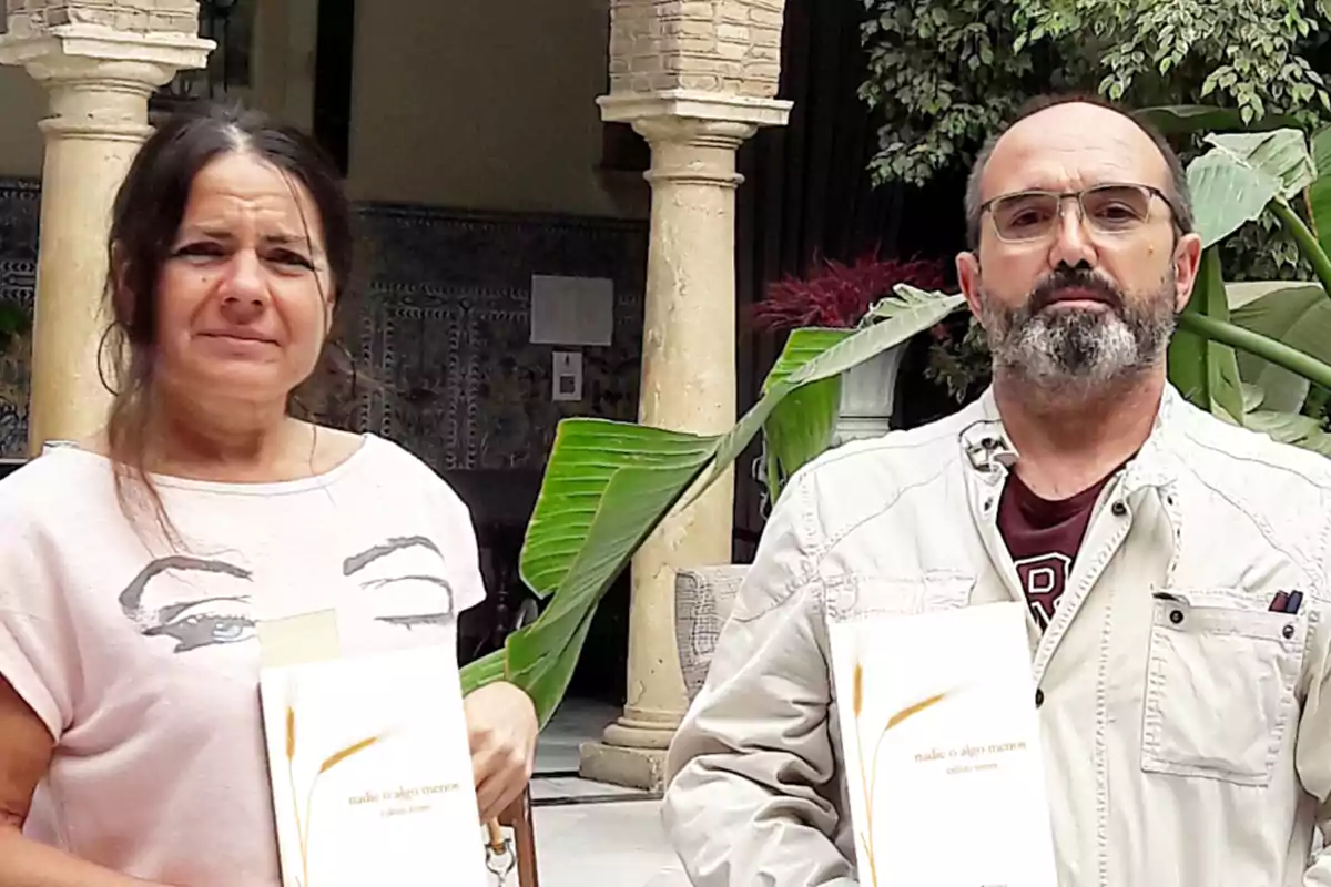 Dos personas posan en un patio con columnas y plantas, cada una sostiene un libro con una portada blanca y detalles dorados.