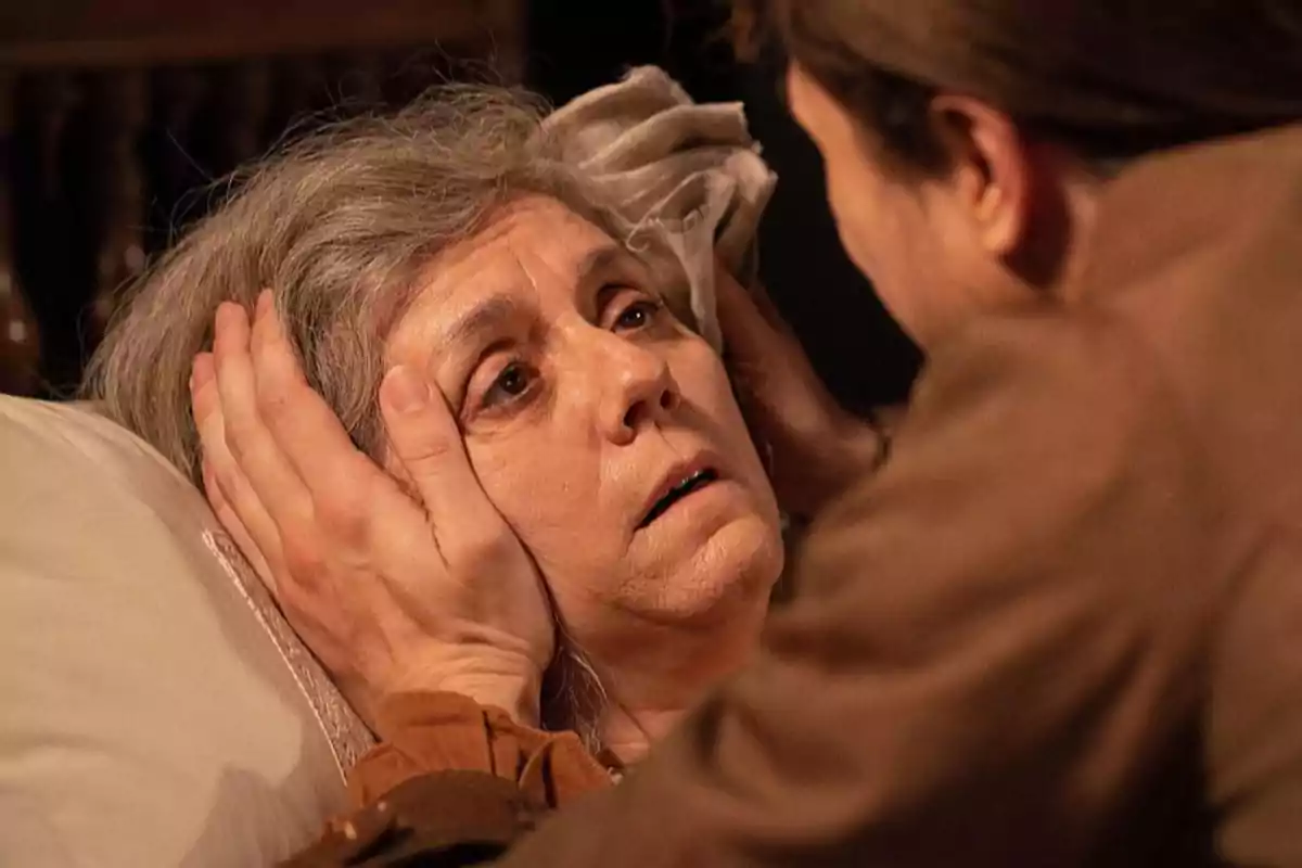 Raimunda recostada en una cama es sostenida por el rostro con ambas manos por otra persona que la mira de cerca mostrando una expresión de preocupación y ternura. Captura de 'Valle Salvaje' Raimunda recostada en una cama es sostenida por el rostro con ambas manos por otra persona que la mira de cerca mostrando una expresión de preocupación y ternura. Captura de 'Valle Salvaje'