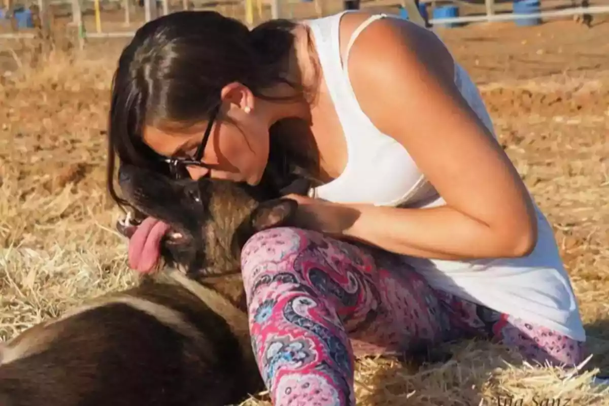Mujer con gafas y camiseta blanca acaricia y besa a un perro que está acostado sobre paja en un campo Mujer con gafas y camiseta blanca acaricia y besa a un perro que está acostado sobre paja en un campo