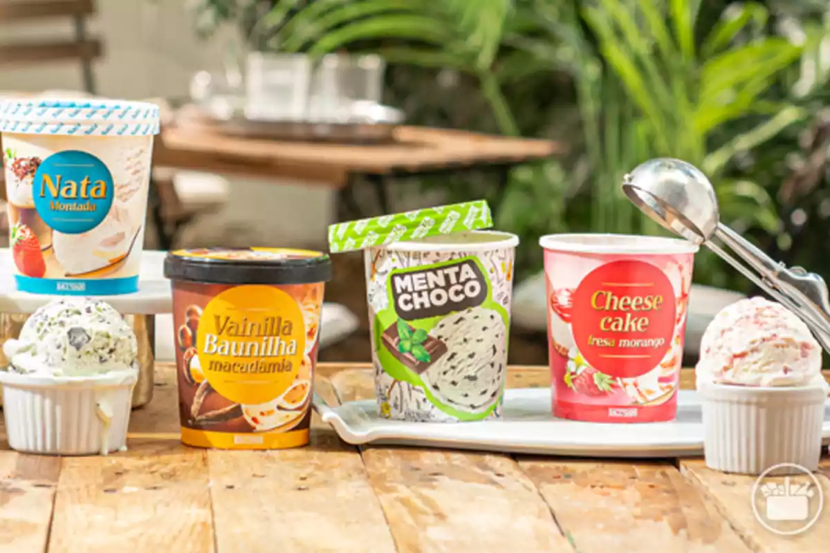 Cuatro envases de helado de diferentes sabores sobre una mesa de madera con dos bolas de helado servidas en pequeños recipientes blancos y una cuchara de helado de metal Cuatro envases de helado de diferentes sabores sobre una mesa de madera con dos bolas de helado servidas en pequeños recipientes blancos y una cuchara de helado de metal