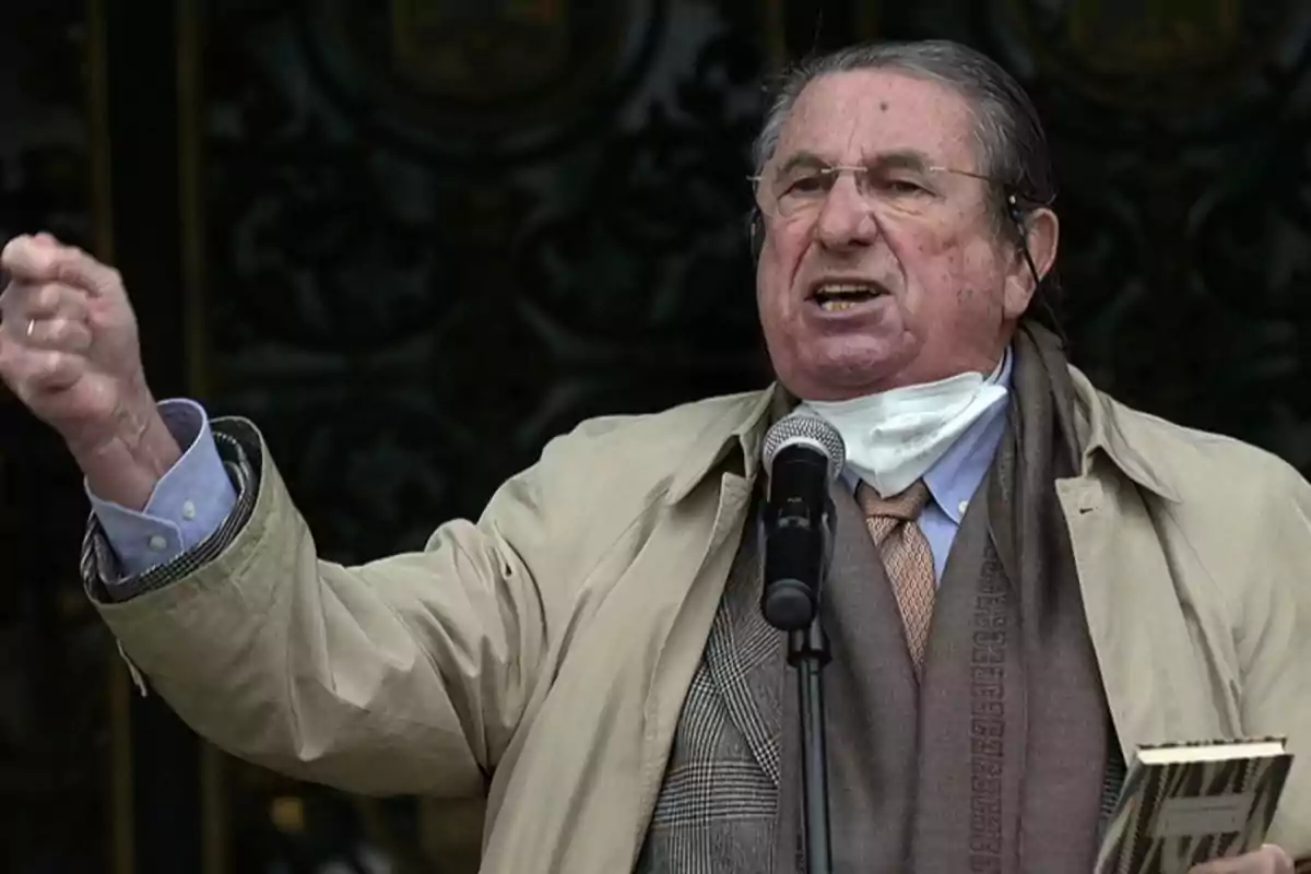 Hombre mayor hablando apasionadamente frente a un micrófono con el puño levantado y una mascarilla bajada al cuello