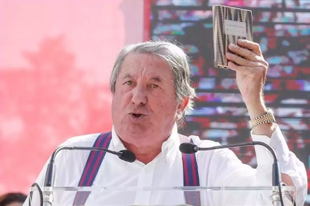 Hombre mayor de cabello canoso hablando en un podio y levantando un libro pequeño con la mano derecha Hombre mayor de cabello canoso hablando en un podio y levantando un libro pequeño con la mano derecha