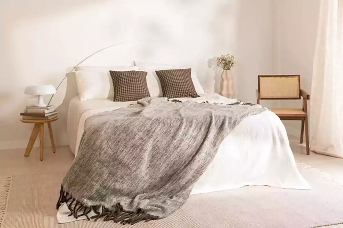 Dormitorio de estilo minimalista con cama vestida en tonos neutros, manta gris, cojines marrones, mesa auxiliar de madera con lámpara blanca y libros, silla de madera con asiento de rejilla y jarrón decorativo con flores secas
