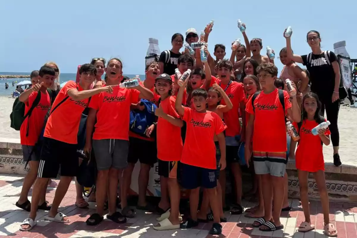 Grupo de niños y adolescentes con camisetas naranjas posando sonrientes en la playa mientras sostienen botellas de bebida en la mano Grupo de niños y adolescentes con camisetas naranjas posando sonrientes en la playa mientras sostienen botellas de bebida en la mano