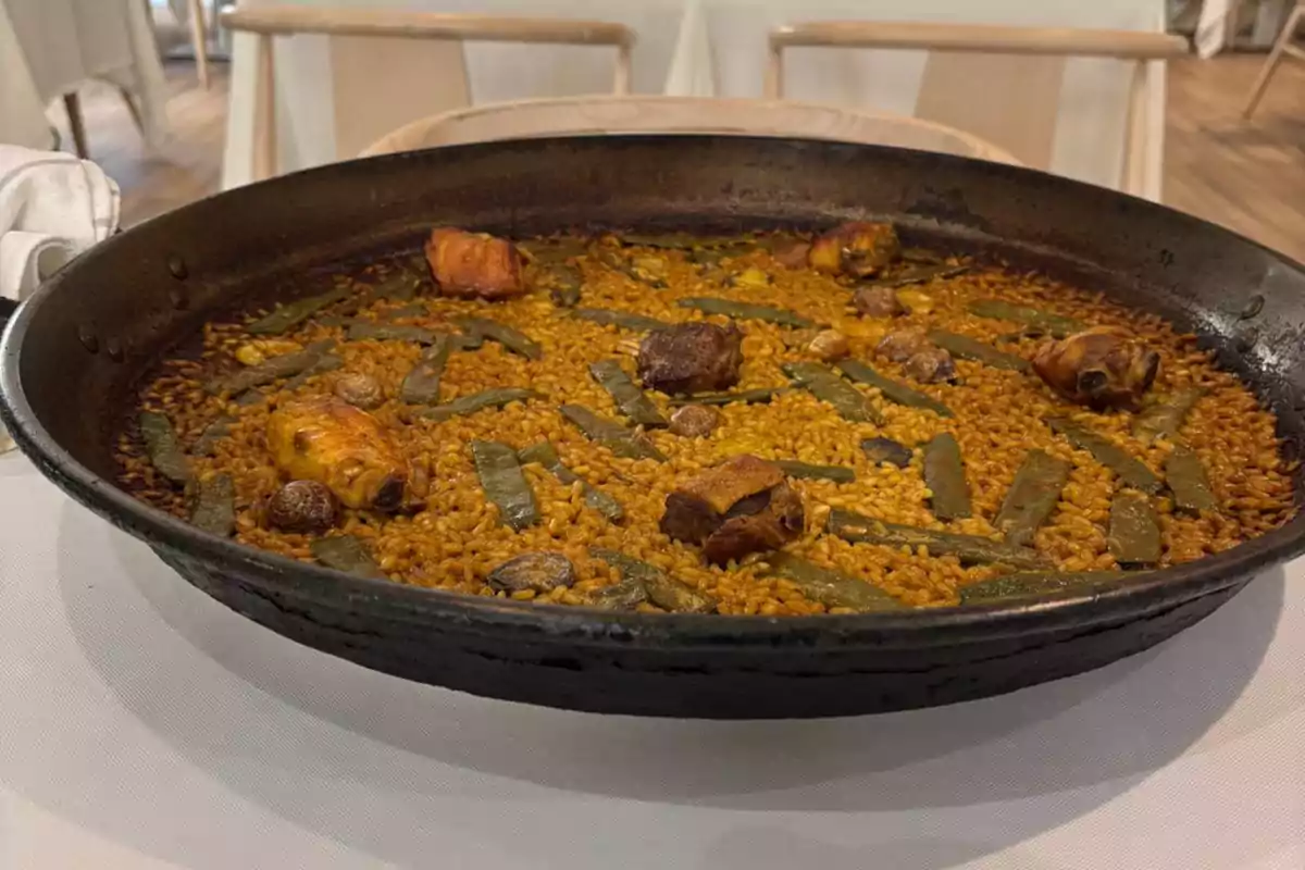 Paella tradicional valenciana servida en una paellera con arroz, trozos de carne, judías verdes y caracoles sobre una mesa blanca Paella tradicional valenciana servida en una paellera con arroz, trozos de carne, judías verdes y caracoles sobre una mesa blanca
