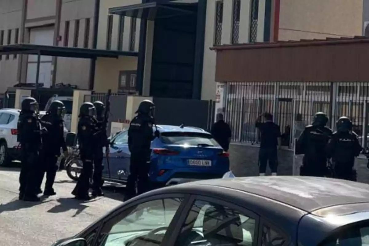 Varios policías con equipo antidisturbios están de pie en una calle frente a un edificio mientras dos personas se encuentran junto a una reja Varios policías con equipo antidisturbios están de pie en una calle frente a un edificio mientras dos personas se encuentran junto a una reja