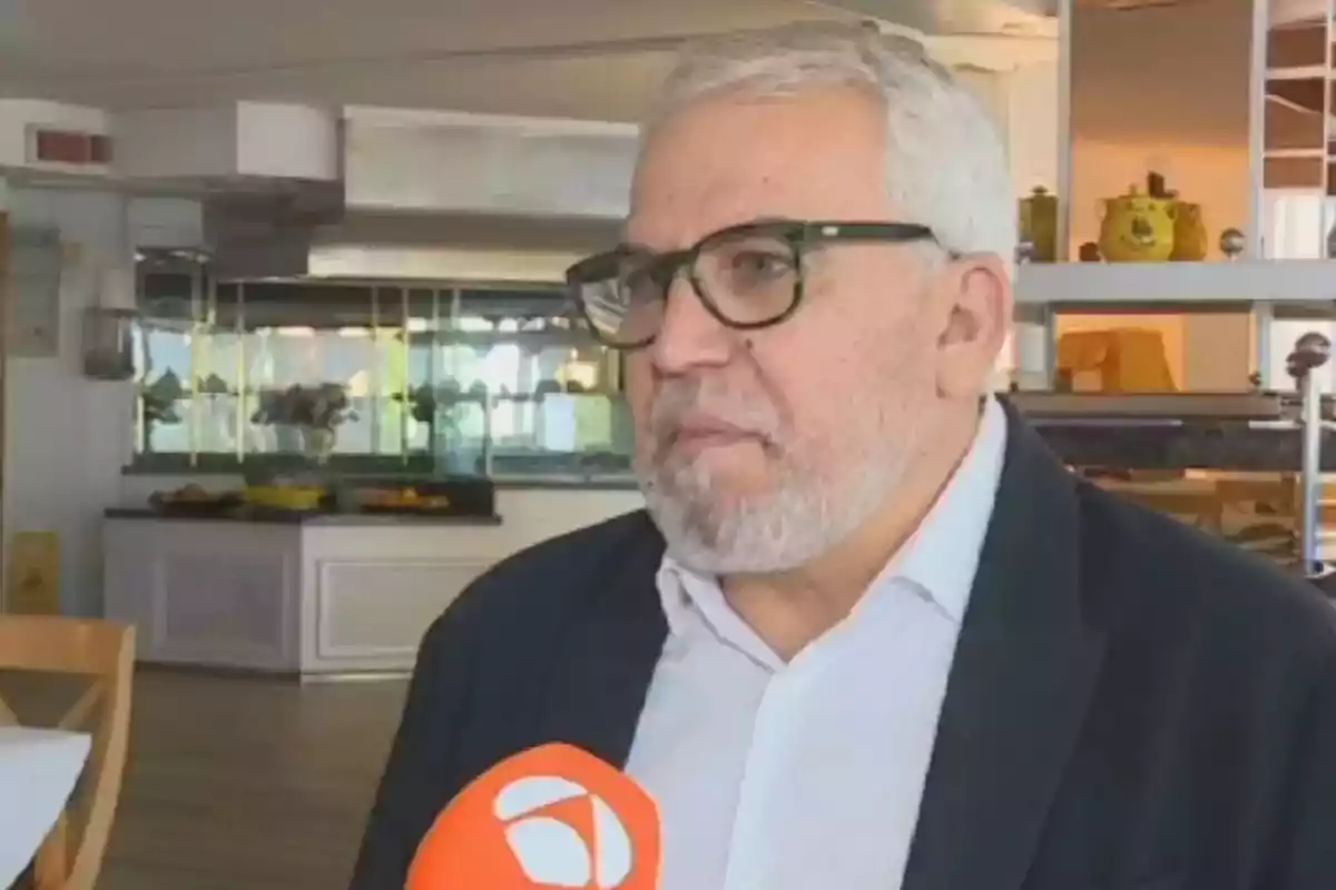 Hombre de cabello canoso y barba, con gafas oscuras, siendo entrevistado en un restaurante mientras sostiene un micrófono naranja Hombre de cabello canoso y barba, con gafas oscuras, siendo entrevistado en un restaurante mientras sostiene un micrófono naranja