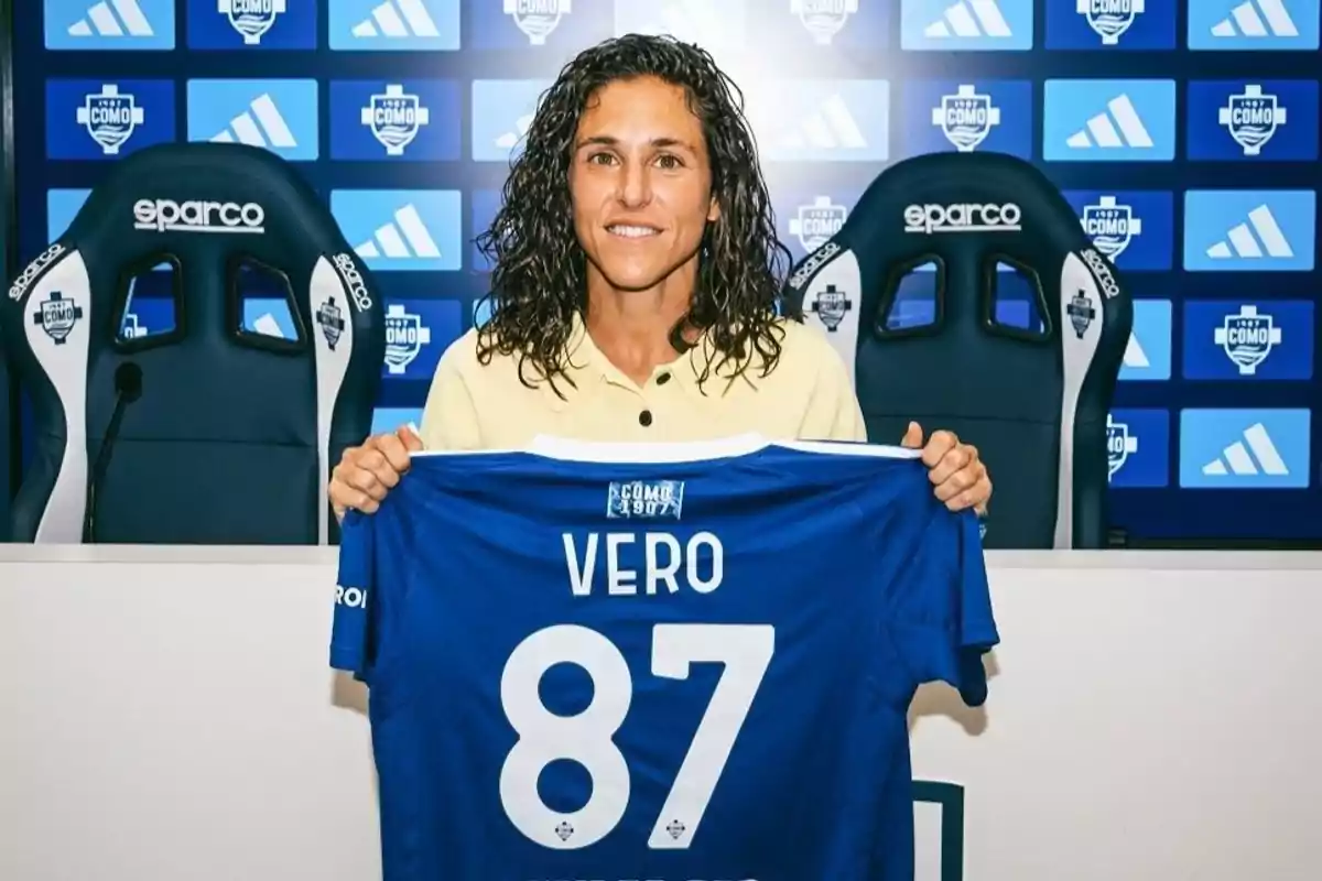 Mujer sonriente con cabello rizado sostiene una camiseta azul con el nombre Vero y el número 87 en una sala de prensa con fondo de logotipos deportivos Mujer sonriente con cabello rizado sostiene una camiseta azul con el nombre Vero y el número 87 en una sala de prensa con fondo de logotipos deportivos