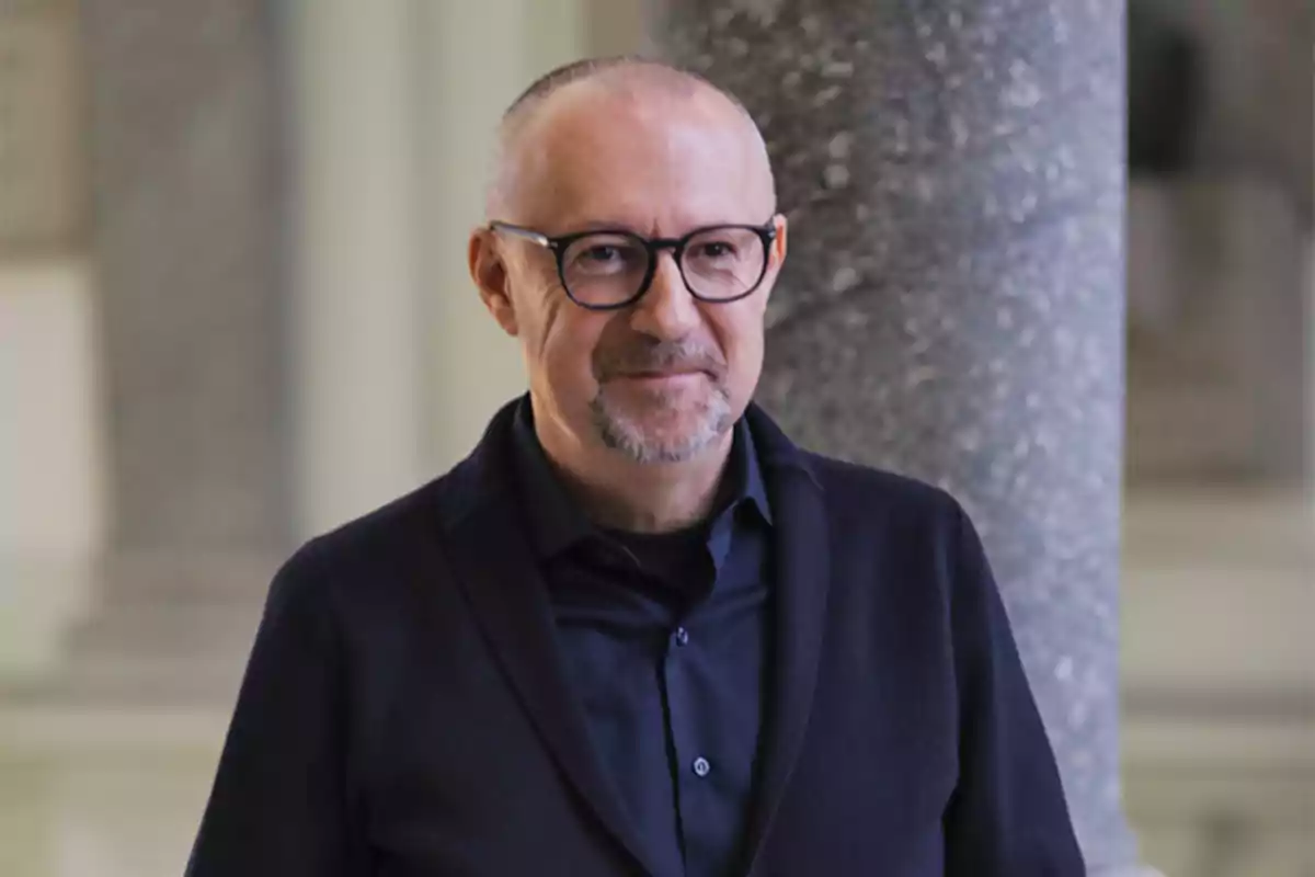 Hombre de mediana edad con gafas y barba corta, vestido con ropa oscura, de pie frente a una columna en un entorno interior elegante. Hombre de mediana edad con gafas y barba corta, vestido con ropa oscura, de pie frente a una columna en un entorno interior elegante.