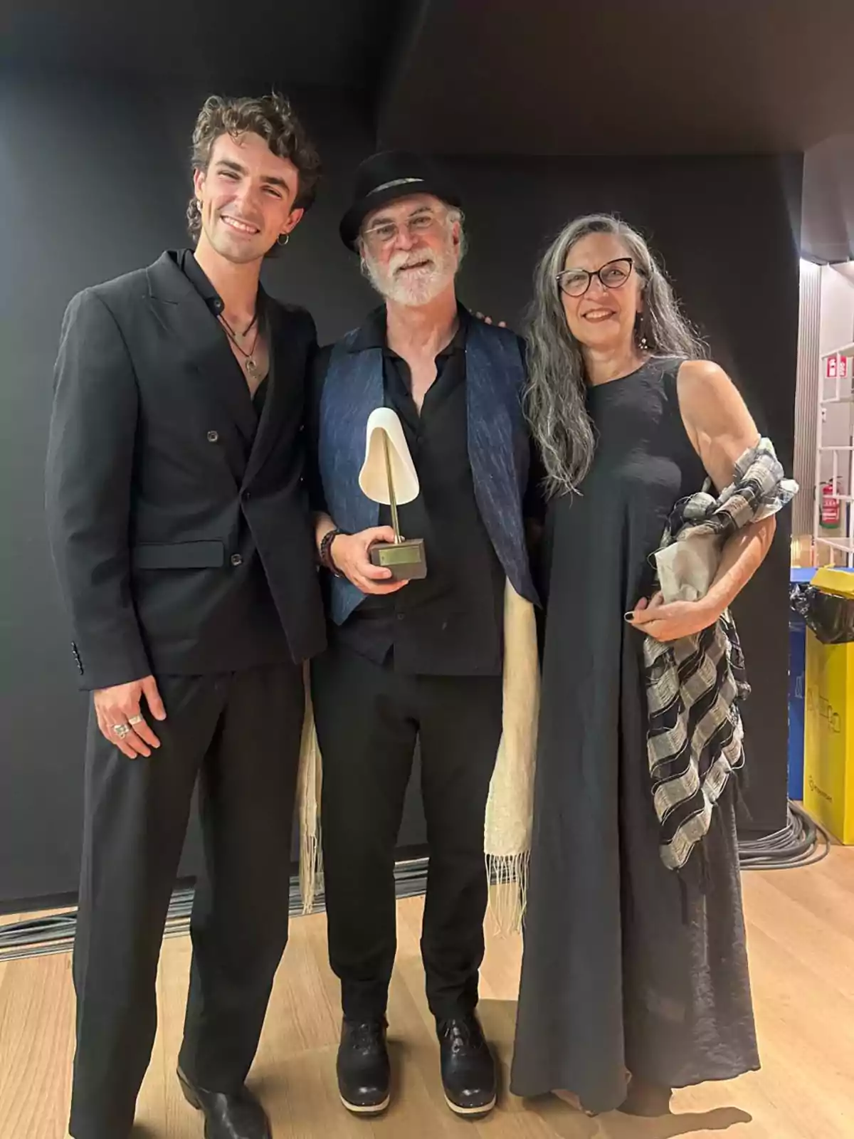 Tres personas posan sonrientes en un interior, el hombre del centro sostiene un trofeo y lleva sombrero, mientras los otros dos lo acompañan vestidos de negro Tres personas posan sonrientes en un interior, el hombre del centro sostiene un trofeo y lleva sombrero, mientras los otros dos lo acompañan vestidos de negro