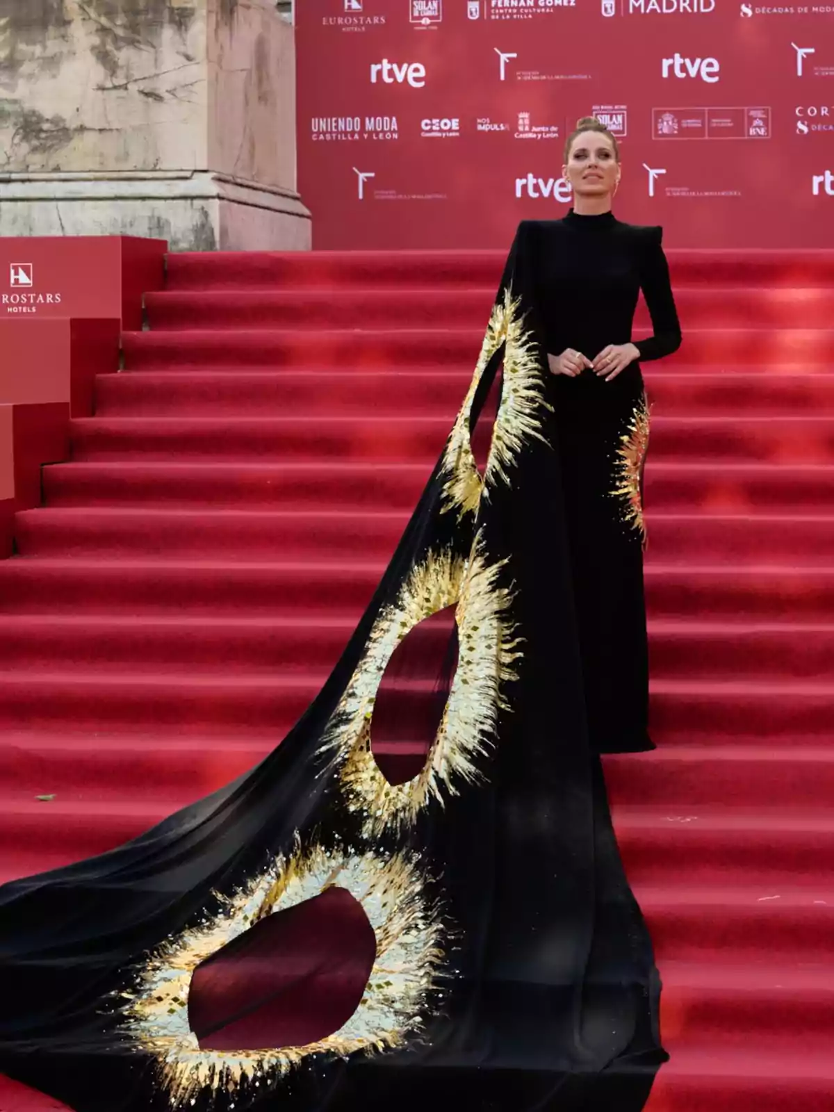 Mujer posando en una alfombra roja con un vestido negro de manga larga y una larga cola decorada con detalles dorados y aberturas circulares Mujer posando en una alfombra roja con un vestido negro de manga larga y una larga cola decorada con detalles dorados y aberturas circulares