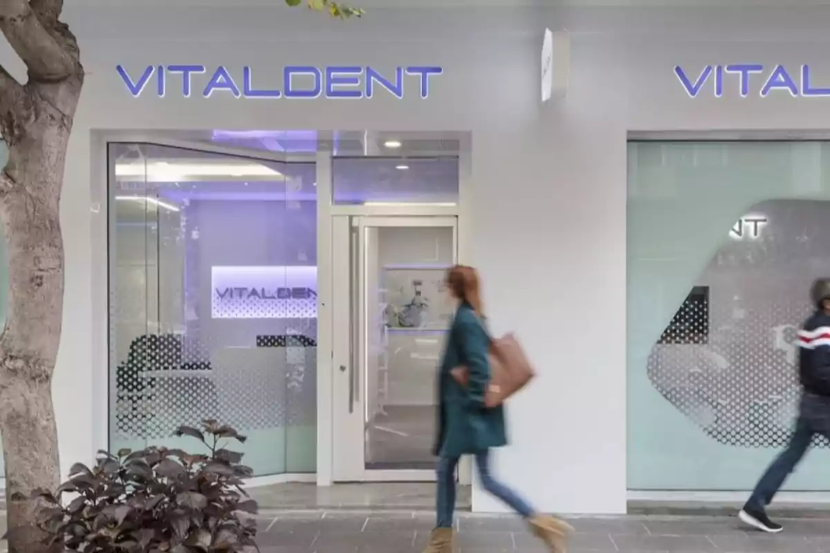 Fachada de una clínica dental Vitaldent con dos personas caminando frente a la entrada Fachada de una clínica dental Vitaldent con dos personas caminando frente a la entrada