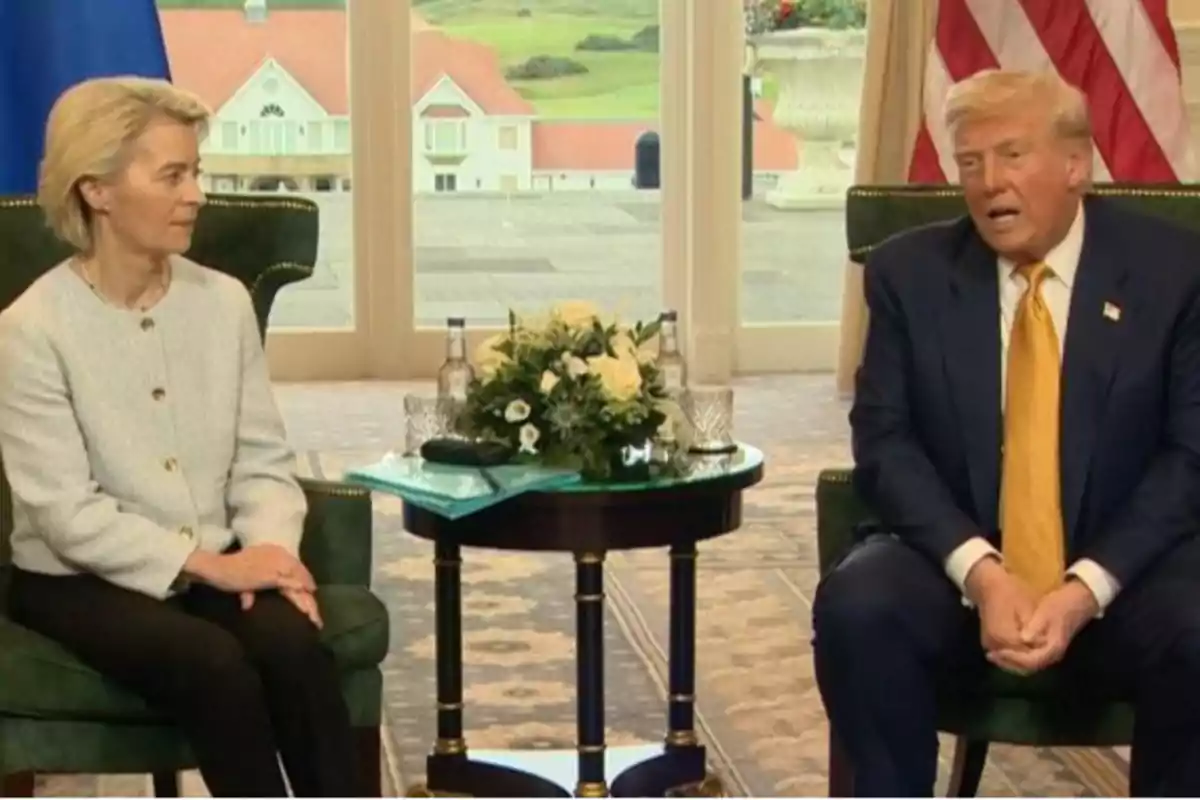 Dos personas sentadas en sillones verdes conversan en una sala elegante con una mesa pequeña entre ellas que tiene flores y botellas de agua al fondo se ven ventanas grandes y una bandera de Estados Unidos