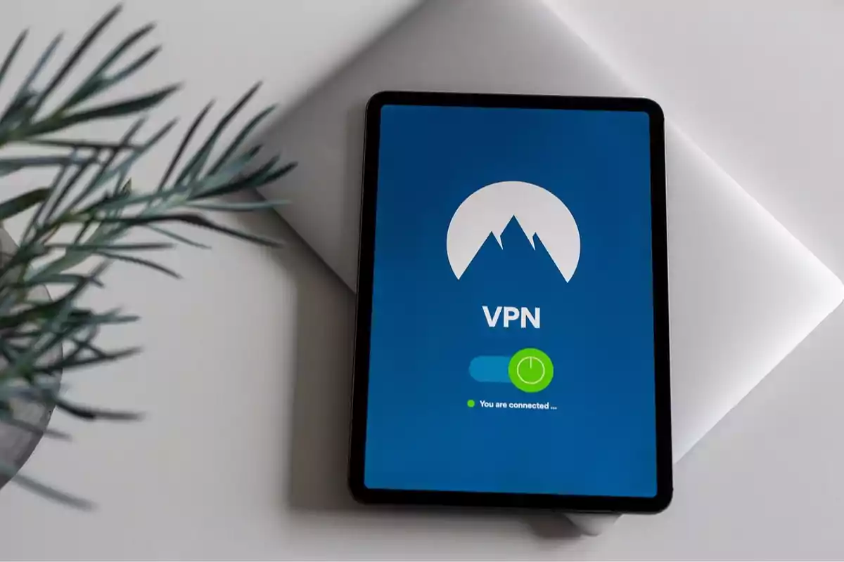 Tablet con pantalla encendida mostrando una aplicación de VPN sobre una laptop plateada en un escritorio blanco junto a una planta