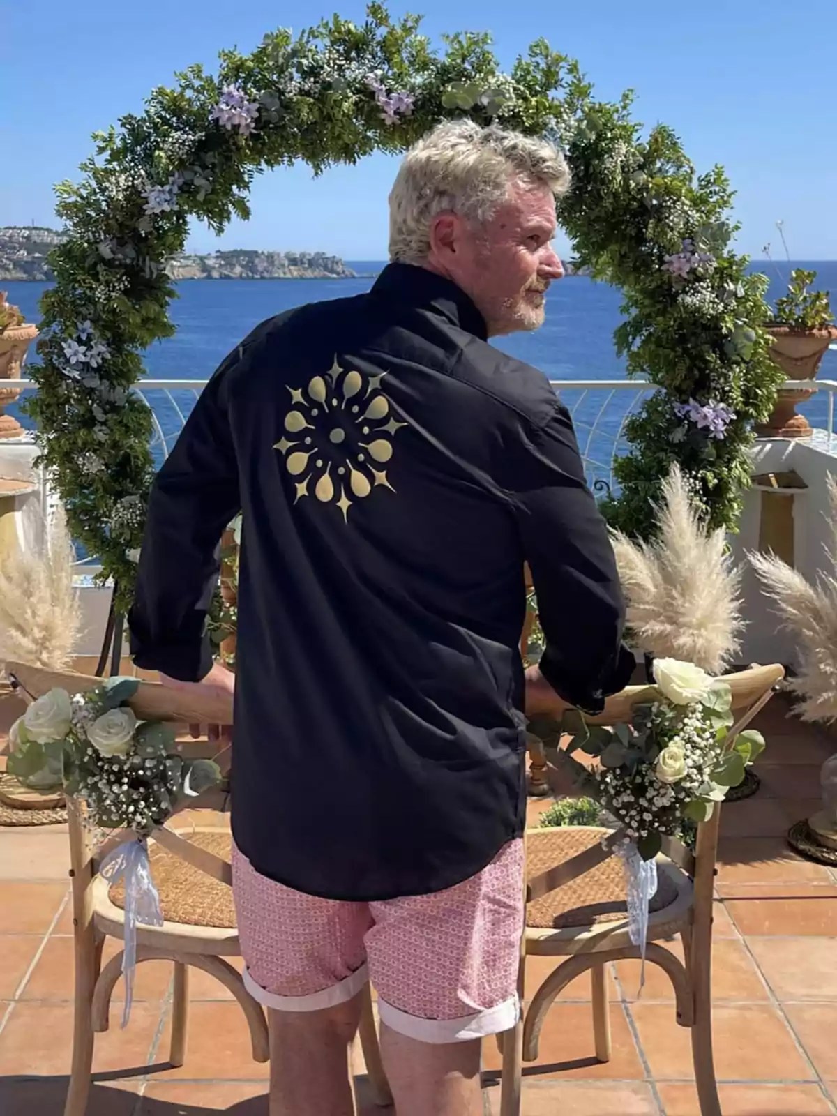 Hombre de cabello rubio con camisa negra y bermuda rosa de espaldas frente a un arco de flores con vista al mar Hombre de cabello rubio con camisa negra y bermuda rosa de espaldas frente a un arco de flores con vista al mar